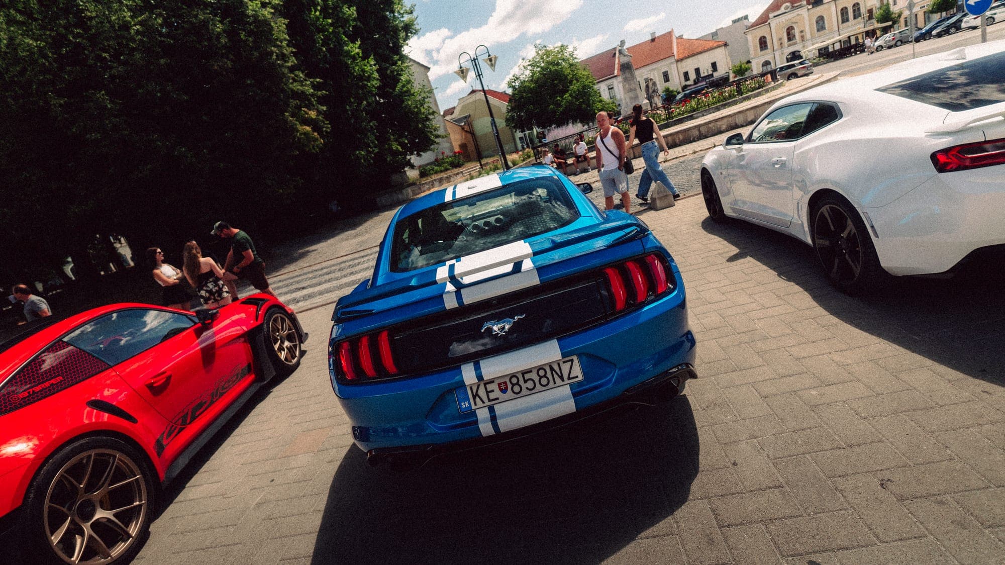 Modrý Mustang s bielymi pruhmi medzi autami na námestí