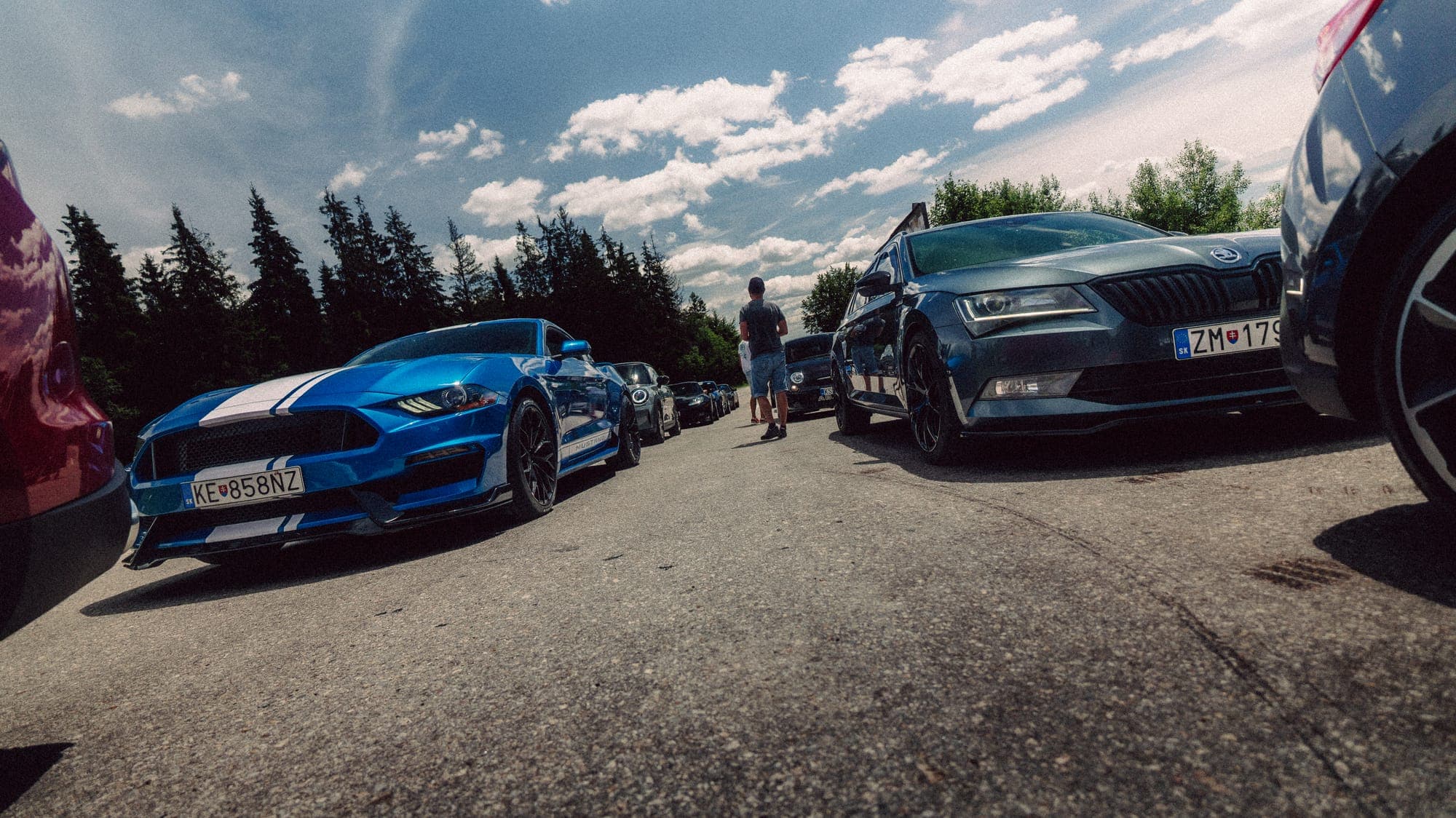 Modrý Mustang a čierne auto na parkovisku pod lesom
